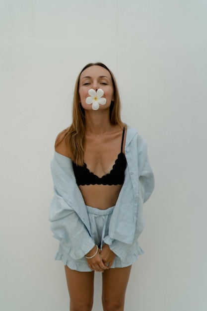 Photo stylish brunette woman in beautiful blue playsuit holding flower and posing over white wall