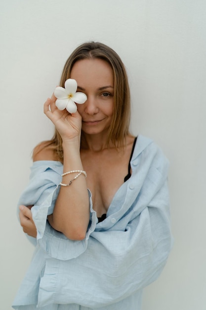 Foto donna castana alla moda in bella tuta blu che tiene fiore e posa sopra il muro bianco