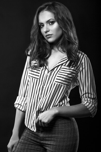 Stylish brunette woman with long curly hair wears striped shirt posing at studio. Black and white portrait