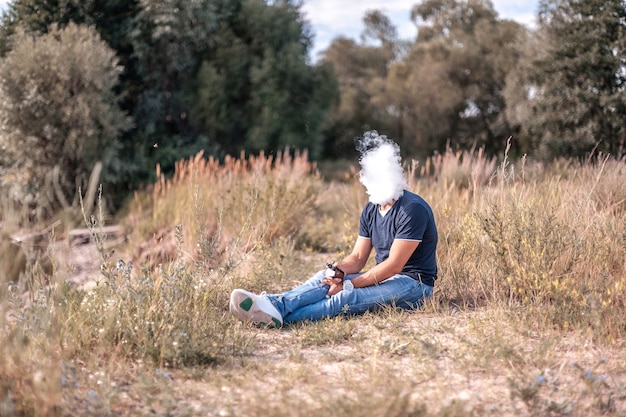 Стильный брутальный курильщик, дующий много дыма, используя электронное дымовое устройство vape на лесной поляне