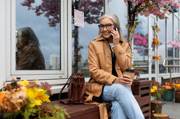 Stylish business elderly woman talking on a mobile phone sitting on a bench next to a flower shop