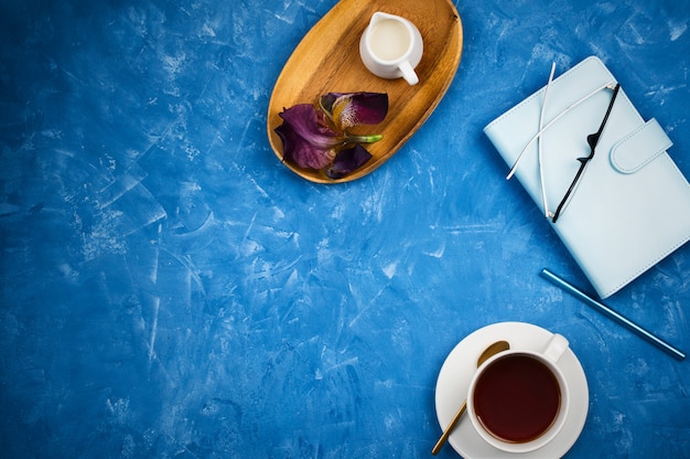 Стильный бизнес-макет с чашкой черного чая, планировщик с очками и ручкой, держатель для молока на деревянном подносе на синем цементном фоне с copyspace