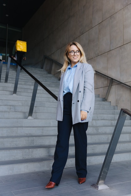 Photo stylish business woman in eyewear and formal outfit standing on stairs near modern building. happy blonde posing on camera outside the office.