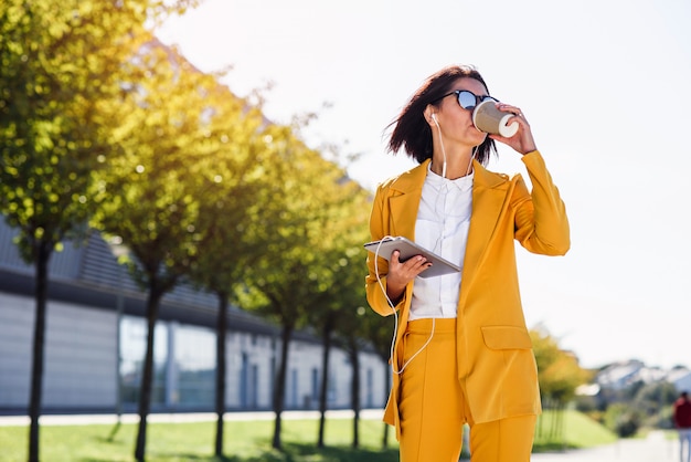 La donna alla moda di affari in vestito giallo con il computer e le cuffie della compressa beve il caffè mentre cammina sul vicolo.