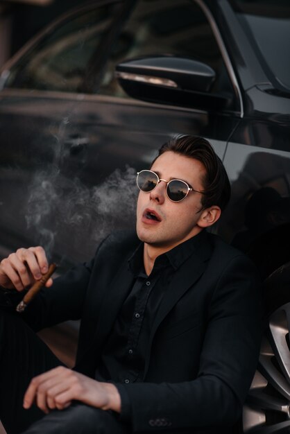 A stylish businessman smokes cigars near a luxury car. Fashion and business
