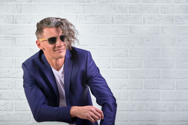 Stylish businessman with curly long hair in sunglasses sitting on chair on white wall