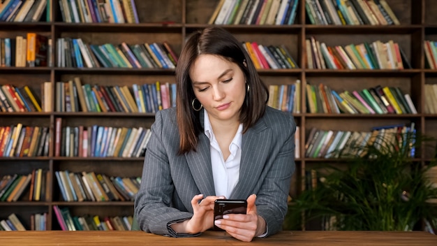 Stylish businesswoman in grey jacket over white blouse types on\
black smartphone and smiles sitting against book racks in library\
hall