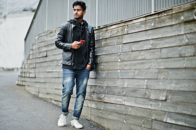 Stylish and casual asian man in black leather jacket, headphones with red mobile phone at hands posed on street