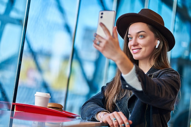 Stylish casual happy smiling hipster brunette female in hat with wireless earphones takes selfie photo using mobile phone during relaxing in restaurant. Modern people lifestyle