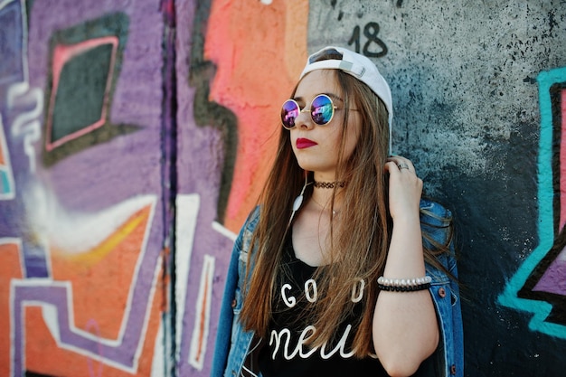 모자 선글라스와 청바지를 입은 세련된 캐주얼 힙스터 소녀는 큰 낙서 벽에 휴대폰 헤드폰으로 음악을 듣습니다.