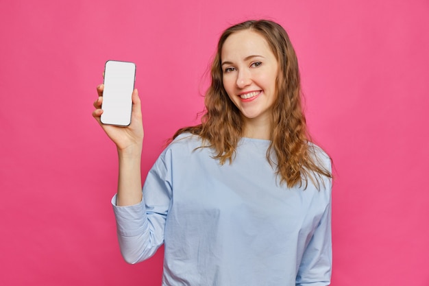 Stylish caucasian girl in a pale blue t-shirt shows smartphone on a pink background.