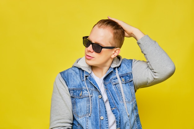 Stylish Caucasian man in a jeans and sunglasses on a yellow background smooths his hair.