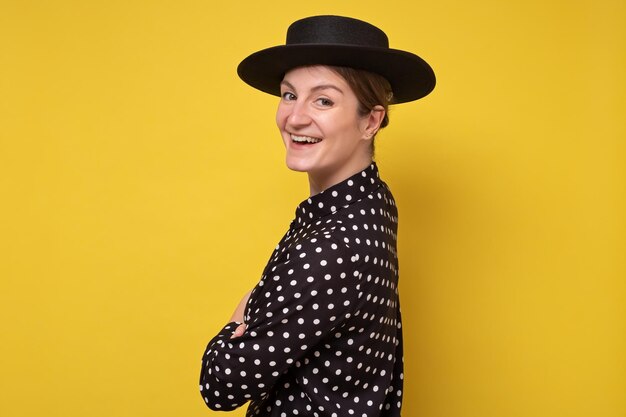 Stylish caucasian woman in black hat looking at camera with confidence. Studio shot on yellow wall.