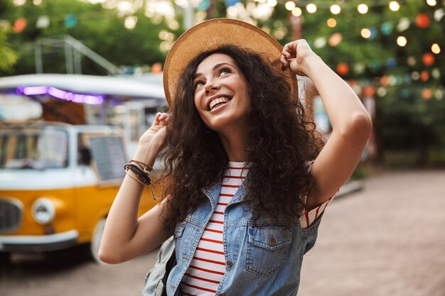 stylish caucasian woman with curly brown hair, laughing and touching her straw hat while rest in hipster spot or modern park