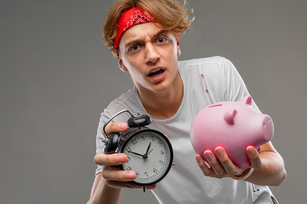 Stylish charming blond guy with a piggy bank and an alarm clock on a gray wall close-up