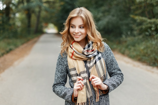 Stylish cheerful European young woman with a cute smile in a fashionable gray coat in a vintage beige checkered scarf walks outdoors on a warm spring day