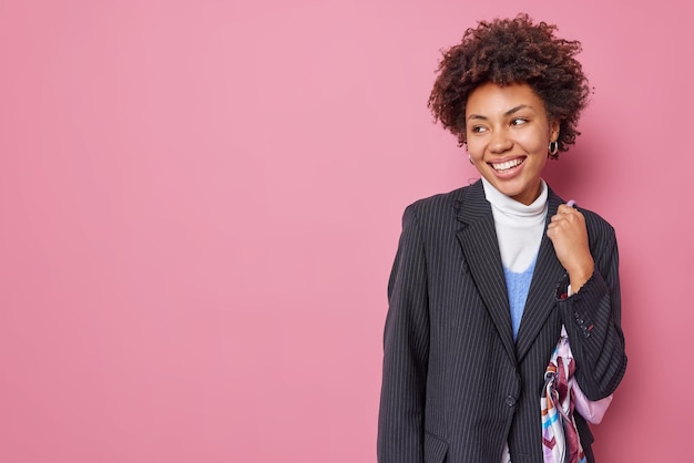 Stylish cheerful woman with natural curly hair dressed in formal black jacket carries bag looks gladfully away sees nice promotion isolated on pink background blank space for your advertising content