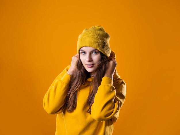 Stylish cheerful young brunette in a yellow hoodie and hat on a yellow background