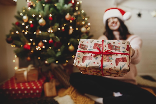 Стильная рождественская подарочная коробка в руках на фоне елки с огнями в праздничной комнате