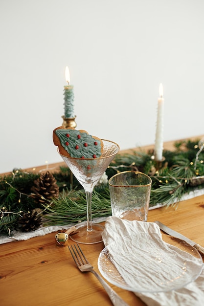 Stylish christmas table setting christmas tree cookie in\
wineglass plate with linen napkin vintage cutlery fir branches with\
golden lights candles on table holiday brunch creative table\
set