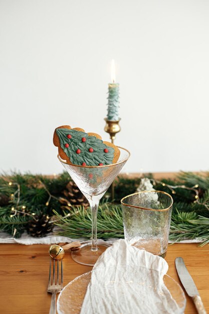 Stylish christmas table setting christmas tree cookie in
wineglass plate with linen napkin vintage cutlery fir branches with
golden lights candles on table holiday brunch creative table
set