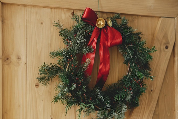 Stylish christmas wreath with red ribbon and golden bells
hanging rustic wooden door winter holiday decor at countryside home
merry christmas and happy holidays