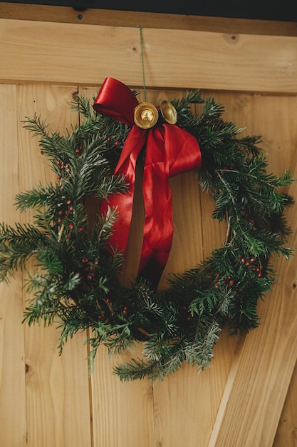 Stylish christmas wreath with red ribbon and golden bells\
hanging rustic wooden door winter holiday decor at countryside home\
merry christmas and happy holidays