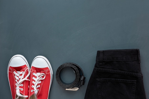 Stylish clothes. Top view of trendy black leather belt, black jeans and youth red sneakers on grey wooden background.