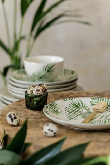 Stylish composition of beautiful plates, cups and kitchen accessories with floral pattern, wooden tray, plants and elegant decoration