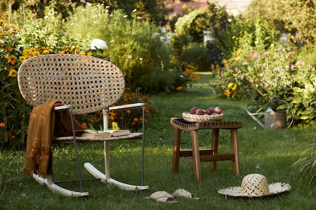 Stylish composition of countryside garden with design rattan armchair, wooden bench, plaid, food, drinks and elegant accessories. A lot of colorful flowers. Summer mood.