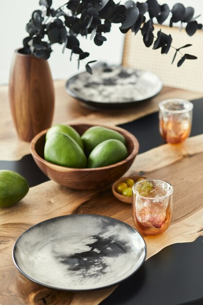 Stylish composition on the design table with elegant plates, glasses, fruits and black eucalyptus flowers in wooden vase. Modern dining room interior.