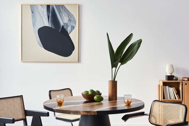 Elegante composizione dell'interno della sala da pranzo con tavolo di design, sedie moderne, decorazione, foglia tropicale in vaso, frutta, quadri astratti e accessori eleganti nell'arredamento della casa