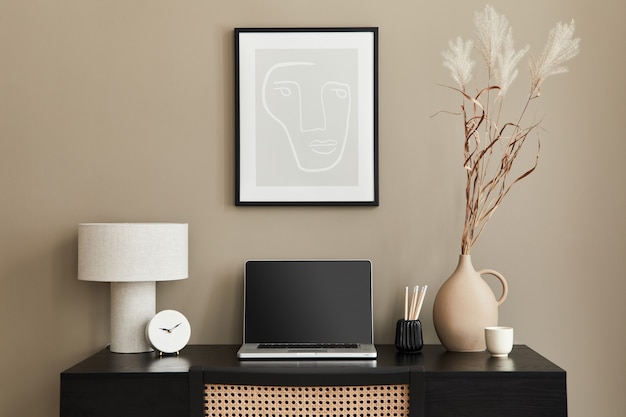 Stylish composition of home office interior with black wooden desk, chair, dried flower in vase, laptop, frame, design table lamp, clock and elegant office accessories.