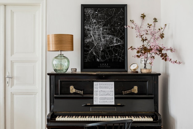 Composizione elegante all'interno del soggiorno con pianoforte nero, mappa poster mock up, fiori secchi, orologio, libro, lampada, parete bianca ed eleganti accessori personali in un arredamento moderno.