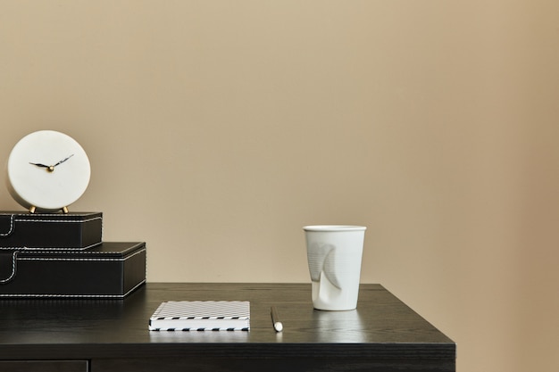 Stylish composition of office interior with black desk, cup of\
coffee, notes, design white clock and desk organizer. template.\
beige wall. copy space.