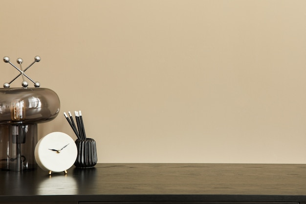 Stylish composition of office interior with black desk, design\
table lamp, white clock and desk organizer. beige wall. copy\
space.