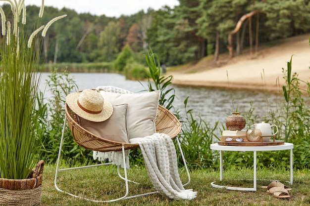 Photo stylish composition of outdoor garden on the lake with design rattan armchair, coffee table, plaid, pillows, drinks and elegant accessories.