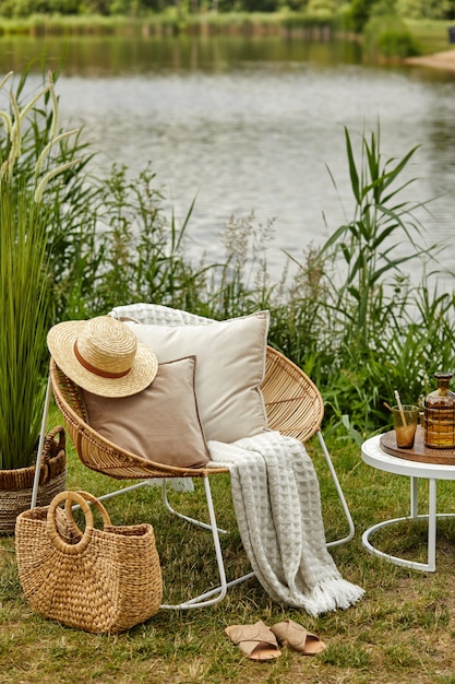디자인 등나무 안락 의자, 커피 테이블, 격자 무늬, 베개, 음료 및 우아한 액세서리와 함께 호수에 야외 정원의 세련된 구성.