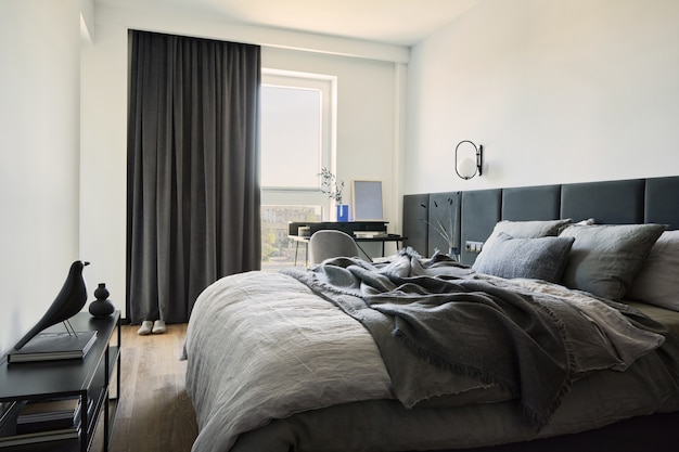 Стильная композиция интерьера небольшой современной спальни. Кровать, креативная лампа и элегантные личные аксессуары. Стены с черными панелями. Панорамные окна. Минималистичная мужская концепция. Шаблон.