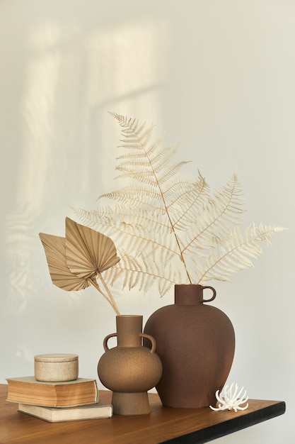 Stylish composition on the wooden table with design vases with\
dried flowers, book and decoration. beautiful shadows on the wall.\
abstract concept in modern interior. template.