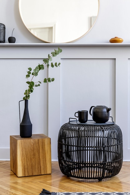 Stylish concept at living room interior with black rattan coffeee table, round mirror, flowers in vase, lantern, shelf, wooden cube and elegant personal accessories in modern home decor.
