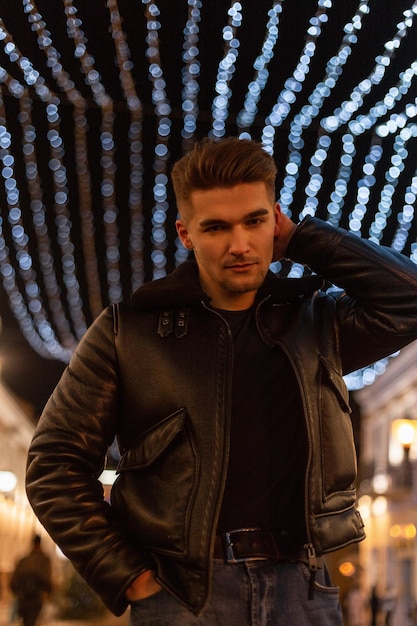 stylish cool young business man model with hairstyle in fashionable black clothes with fashion leather jacket walking in the city with lights at night