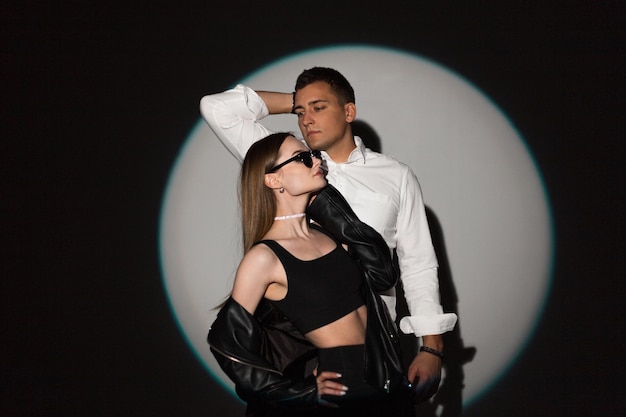 Stylish cool young couple beautiful girl with sunglasses and stylish handsome man model in fashionable casual clothes in studio on a dark background at a party