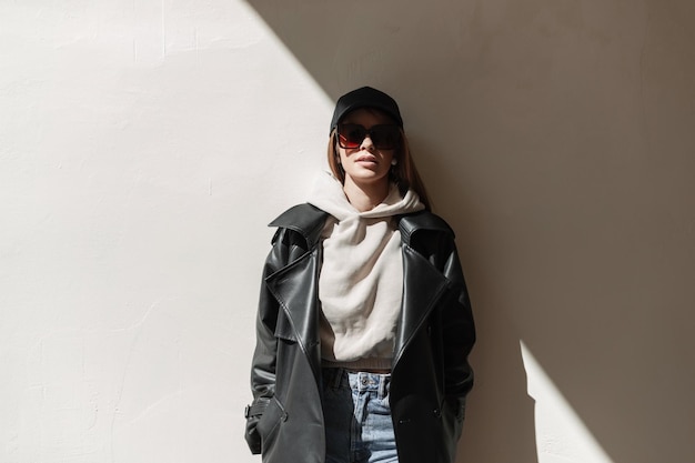 stylish cool young hipster woman model with a black cap and trendy sunglasses in a fashion black leather coat jeans with a hoodie standing near a wall with sunlight and shade urban female style look
