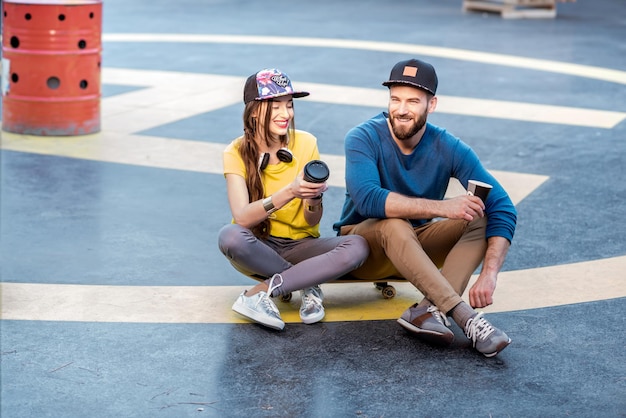 Stylish couple in caps sitting together with coffee cups on the skateboard outdoors on the helipad ground