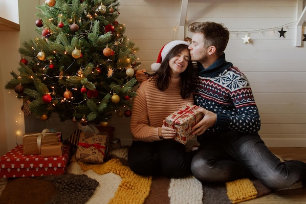 Stylish couple exchanging christmas gifts under christmas tree with presents and lights