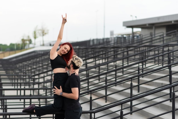 Stylish couple in love in black clothes walks in the park and hugs Lovely couple of hipsters hugging on the background of the stairs Emotional girl with hands up