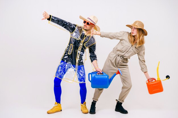 Stylish couple of man and woman in straw hats posing with watering pot over white wall