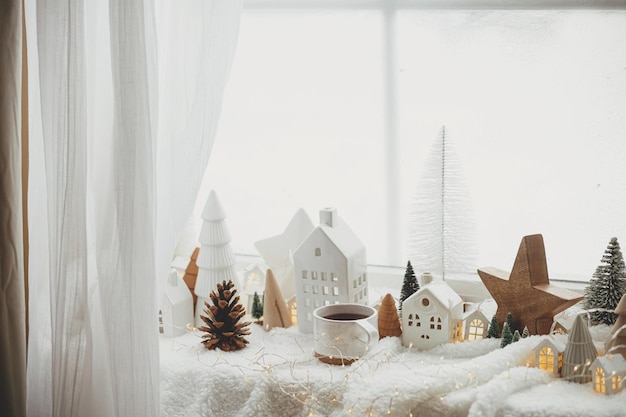 モダンなクリスマスの家とスタイリッシュなお茶のカップ松ぼっくりの木の星と木の装飾金色のライトが窓辺の暖かい毛布の上に居心地の良いクリスマス北欧スタイルのクリスマスの背景
