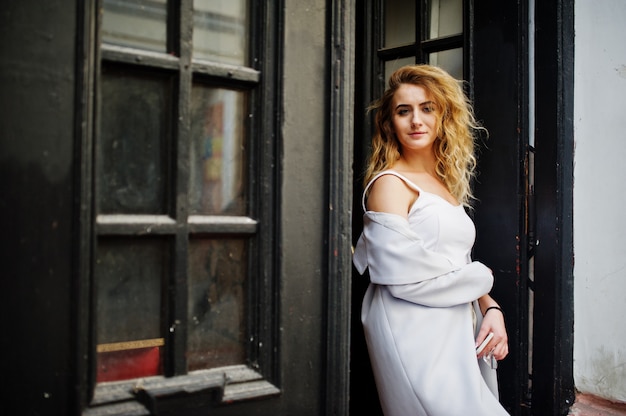 Stylish curly blonde model girl wear on white posing against old wooden door.